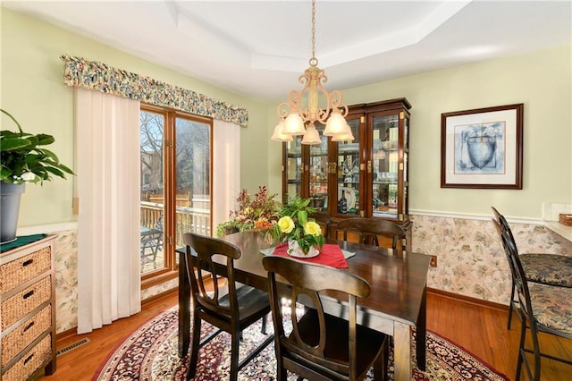 dining area with a wainscoted wall, wood finished floors, visible vents, a tray ceiling, and wallpapered walls