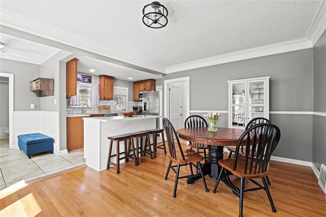 dining room with a wainscoted wall, a textured ceiling, crown molding, and light wood-style floors