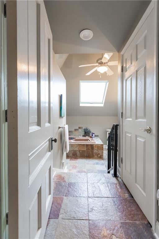 interior space featuring lofted ceiling with skylight, stone tile flooring, a garden tub, and a ceiling fan