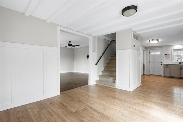 unfurnished living room featuring a decorative wall, visible vents, stairway, light wood-type flooring, and beamed ceiling