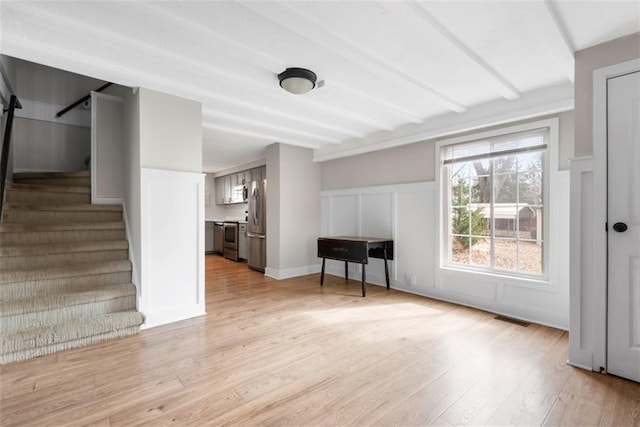 unfurnished living room with light wood-style flooring, a decorative wall, visible vents, stairway, and beamed ceiling