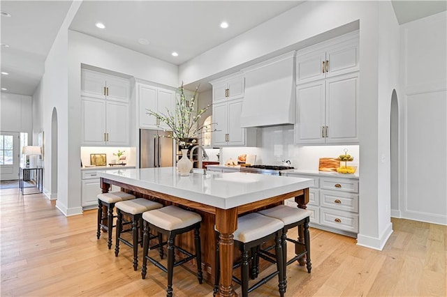 kitchen featuring arched walkways, custom exhaust hood, light countertops, an island with sink, and light wood-type flooring