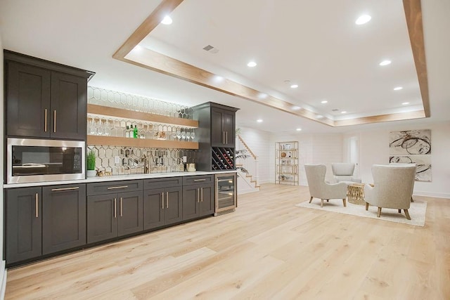 bar with wine cooler, light wood-type flooring, wet bar, stainless steel microwave, and a raised ceiling