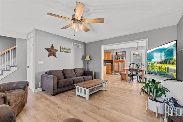 living room featuring a ceiling fan, visible vents, baseboards, light wood-style flooring, and stairs