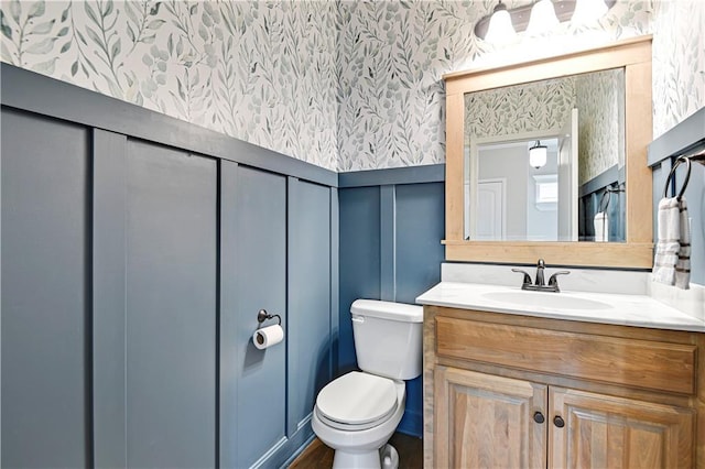 half bathroom with a decorative wall, toilet, wallpapered walls, and vanity