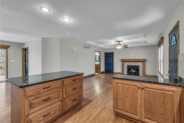 kitchen with visible vents, dark stone counters, a lit fireplace, a tray ceiling, and light wood-style floors