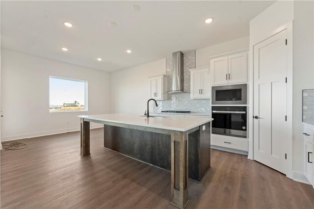 kitchen with a center island with sink, stainless steel microwave, oven, wall chimney range hood, and a sink