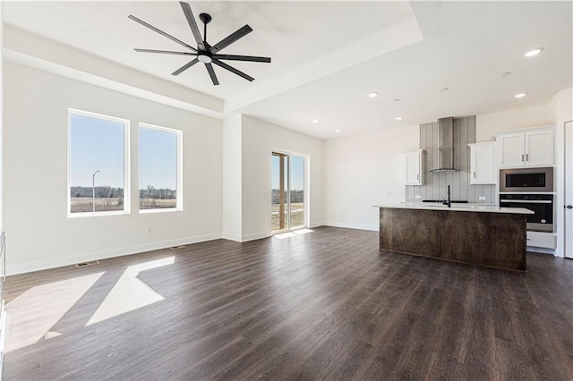 kitchen with dark wood-style flooring, stainless steel microwave, a kitchen island with sink, wall chimney range hood, and oven