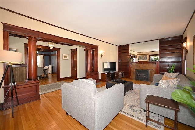 living room featuring baseboards, ornate columns, light wood-style flooring, ornamental molding, and a brick fireplace