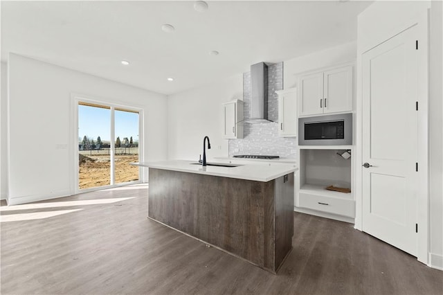 kitchen featuring dark wood-style floors, stainless steel microwave, a kitchen island with sink, a sink, and wall chimney range hood