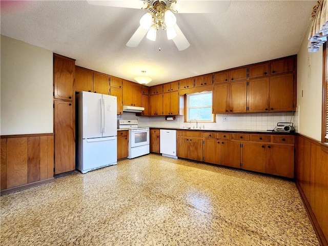 kitchen with under cabinet range hood, a wainscoted wall, white appliances, and brown cabinetry