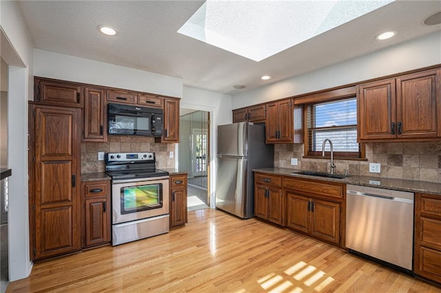 kitchen featuring stainless steel appliances, a sink, backsplash, light wood finished floors, and dark stone countertops