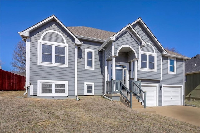 raised ranch with driveway, roof with shingles, an attached garage, and fence