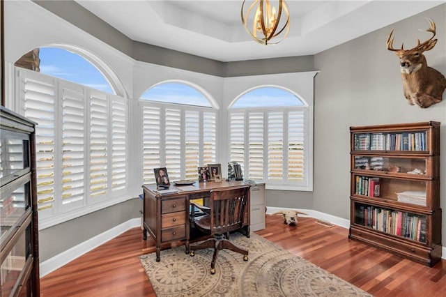 home office featuring a raised ceiling, a notable chandelier, baseboards, and wood finished floors