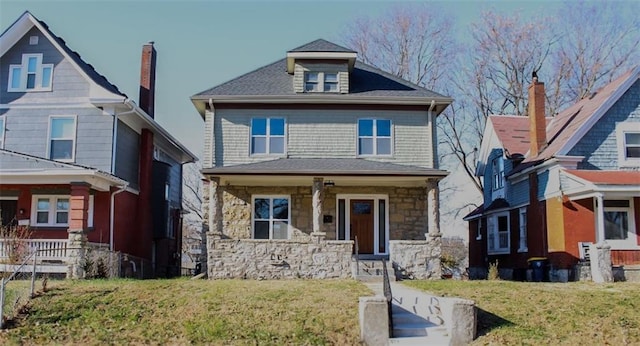 american foursquare style home featuring stone siding, roof with shingles, covered porch, and a front yard