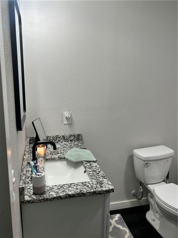 half bath featuring toilet, a textured wall, vanity, and baseboards