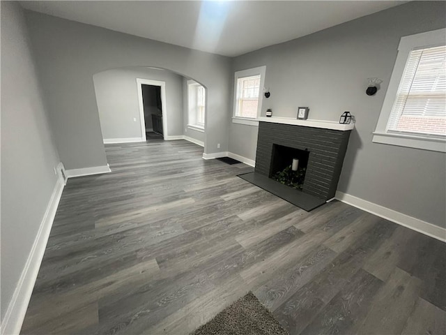 unfurnished living room with arched walkways, a brick fireplace, dark wood finished floors, and baseboards