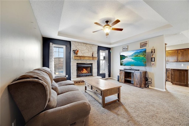 living room featuring a textured ceiling, a stone fireplace, a ceiling fan, baseboards, and a tray ceiling