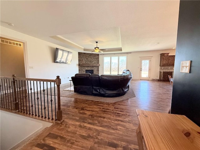 living room with visible vents, a ceiling fan, a tray ceiling, wood finished floors, and a stone fireplace