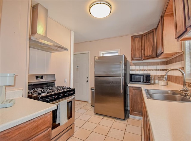 kitchen featuring light tile patterned floors, brown cabinetry, a sink, appliances with stainless steel finishes, and wall chimney range hood