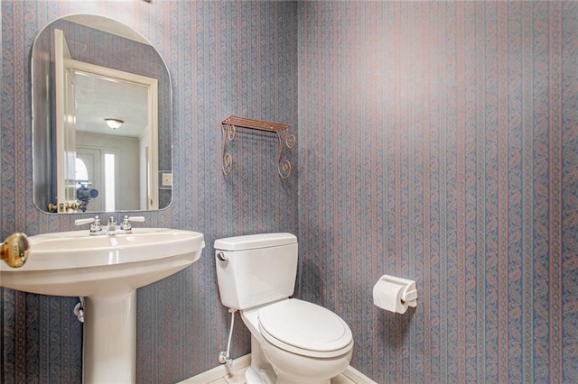 bathroom with a sink, toilet, baseboards, and wallpapered walls