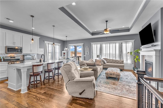 living area featuring ceiling fan, recessed lighting, wood finished floors, a tray ceiling, and a glass covered fireplace