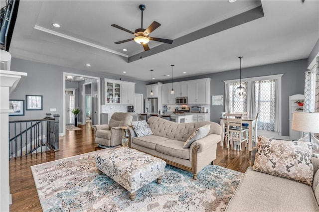 living room with recessed lighting, ceiling fan with notable chandelier, baseboards, a tray ceiling, and dark wood finished floors