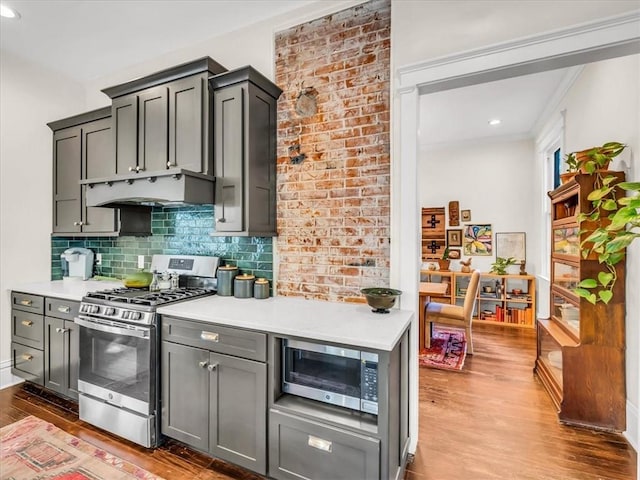 kitchen featuring under cabinet range hood, stainless steel appliances, wood finished floors, light countertops, and tasteful backsplash