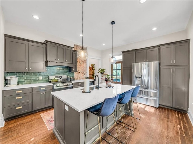 kitchen with wood finished floors, hanging light fixtures, appliances with stainless steel finishes, backsplash, and a center island with sink