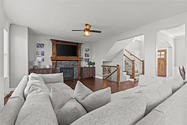 living area featuring ceiling fan, dark wood finished floors, a stone fireplace, stairs, and baseboards