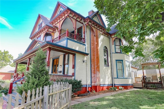 view of front facade with a balcony, covered porch, fence, a chimney, and a front yard