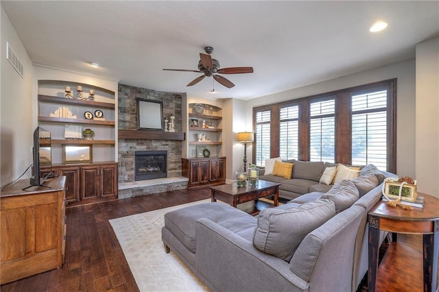 living room featuring built in shelves, a stone fireplace, dark wood-style flooring, visible vents, and a ceiling fan