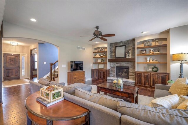 living room featuring built in features, arched walkways, dark wood-style flooring, visible vents, and a stone fireplace