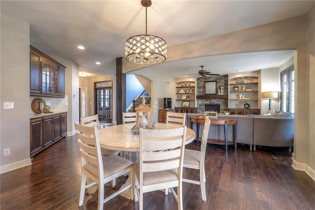 dining room featuring ceiling fan with notable chandelier, baseboards, dark wood-style flooring, and a stone fireplace