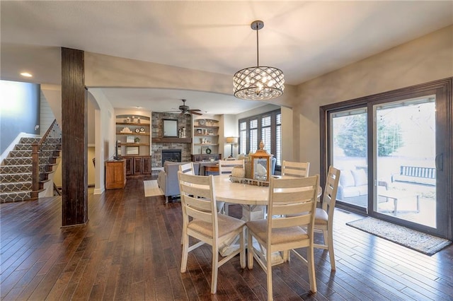 dining area with a stone fireplace, a ceiling fan, built in features, stairway, and dark wood finished floors