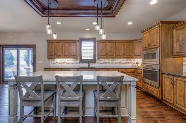 kitchen with brown cabinetry, a raised ceiling, dark wood-style floors, appliances with stainless steel finishes, and a sink