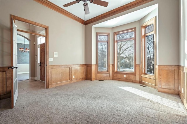 unfurnished room featuring a wainscoted wall, ceiling fan with notable chandelier, carpet flooring, and crown molding