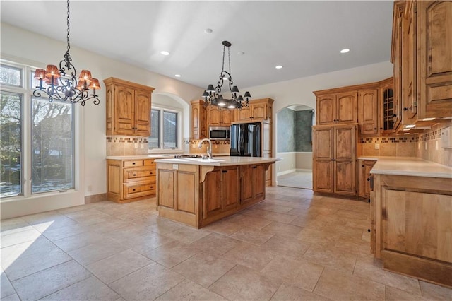 kitchen with arched walkways, black refrigerator, light countertops, stainless steel microwave, and an island with sink