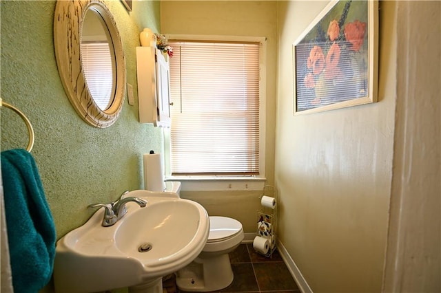 half bath with baseboards, a sink, tile patterned floors, toilet, and a textured wall