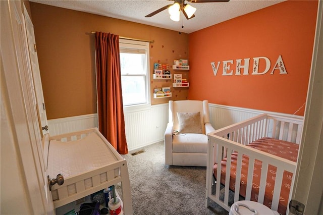 bedroom with visible vents, a wainscoted wall, carpet floors, a textured ceiling, and a nursery area