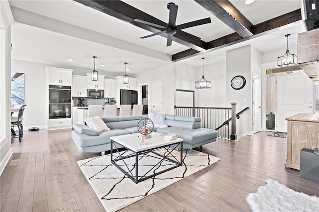 living area featuring ceiling fan with notable chandelier, light wood finished floors, beam ceiling, and baseboards