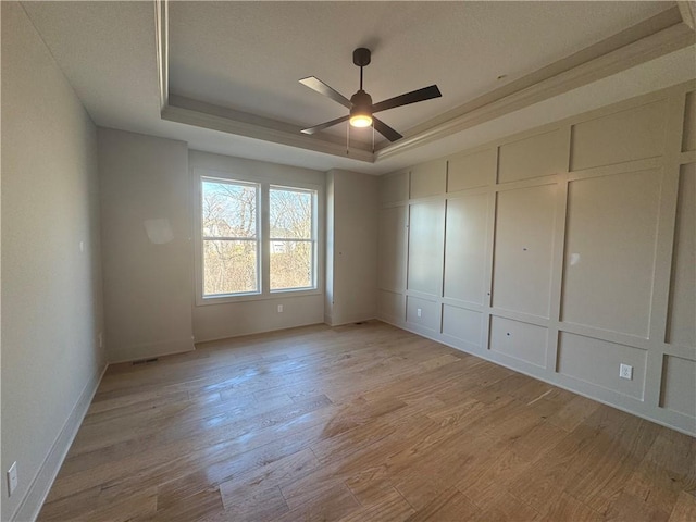 empty room featuring light wood-type flooring, a raised ceiling, a ceiling fan, and a decorative wall