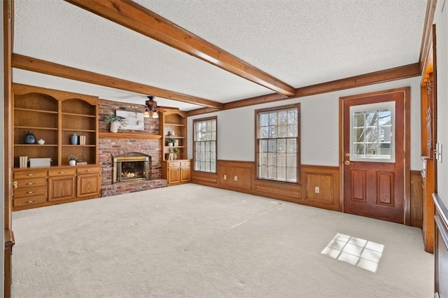 unfurnished living room with carpet, beam ceiling, a fireplace, wainscoting, and a textured ceiling