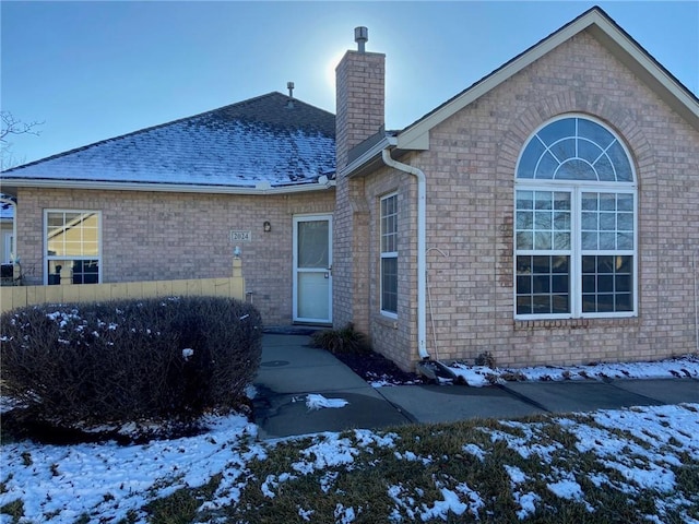 snow covered house with brick siding, a chimney, and a shingled roof