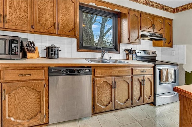 kitchen with stainless steel appliances, light countertops, brown cabinetry, a sink, and under cabinet range hood