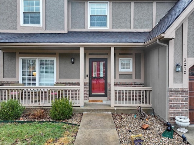 view of exterior entry featuring brick siding, covered porch, roof with shingles, and stucco siding