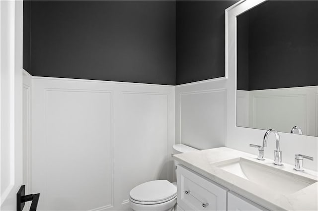 half bath with wainscoting, a decorative wall, vanity, and toilet