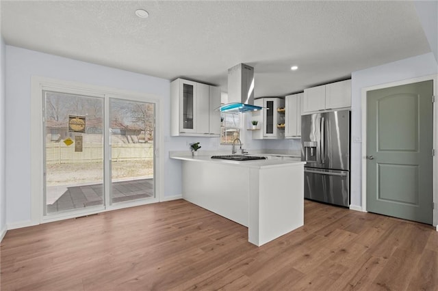 kitchen featuring a peninsula, island exhaust hood, stainless steel fridge with ice dispenser, light countertops, and glass insert cabinets