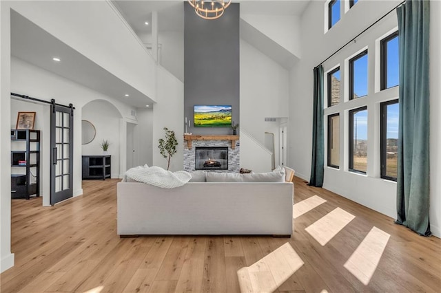 living area featuring light wood-style flooring, arched walkways, a stone fireplace, a towering ceiling, and a barn door