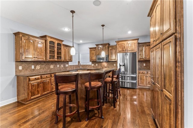 kitchen with appliances with stainless steel finishes, a breakfast bar area, brown cabinetry, glass insert cabinets, and dark wood-style flooring